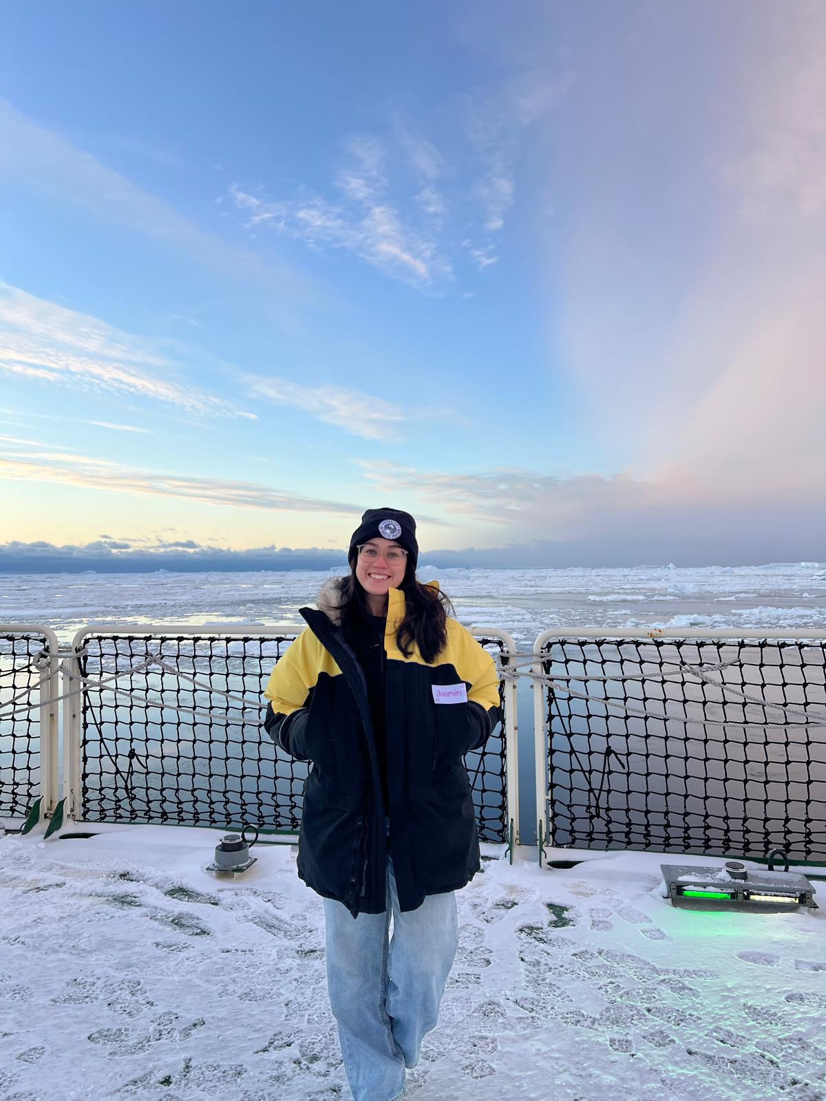 Marine Geoscientist Jasmin Wells stands on the helideck aboard the RSV Nuyina. The deck is coated in snow with ice bergs in the ocean behind her.