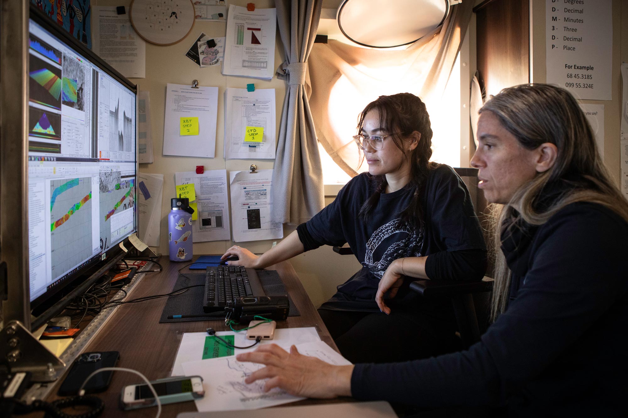 Two women at a desk in a ship's cabin examining complex seabed data visualisations on a computer screen