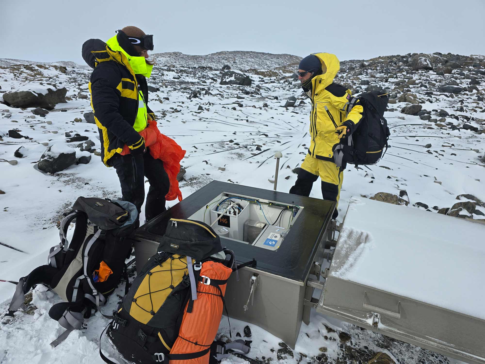 Two men in brightly coloured survival gear stand in a rocky snow-covered field next to a metal box that contains scientific equipment
