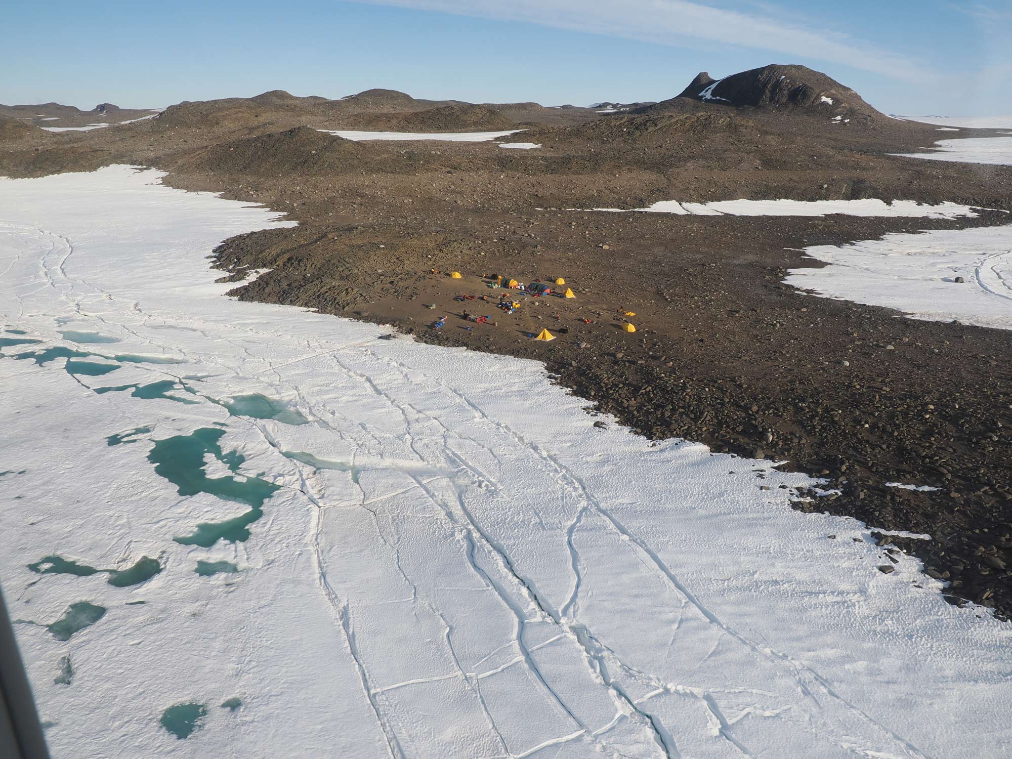 An aerial shot of bare rocky terrain surrounded with ice. A small collection of bright yellow huts and tents are visible in the centre of the shot