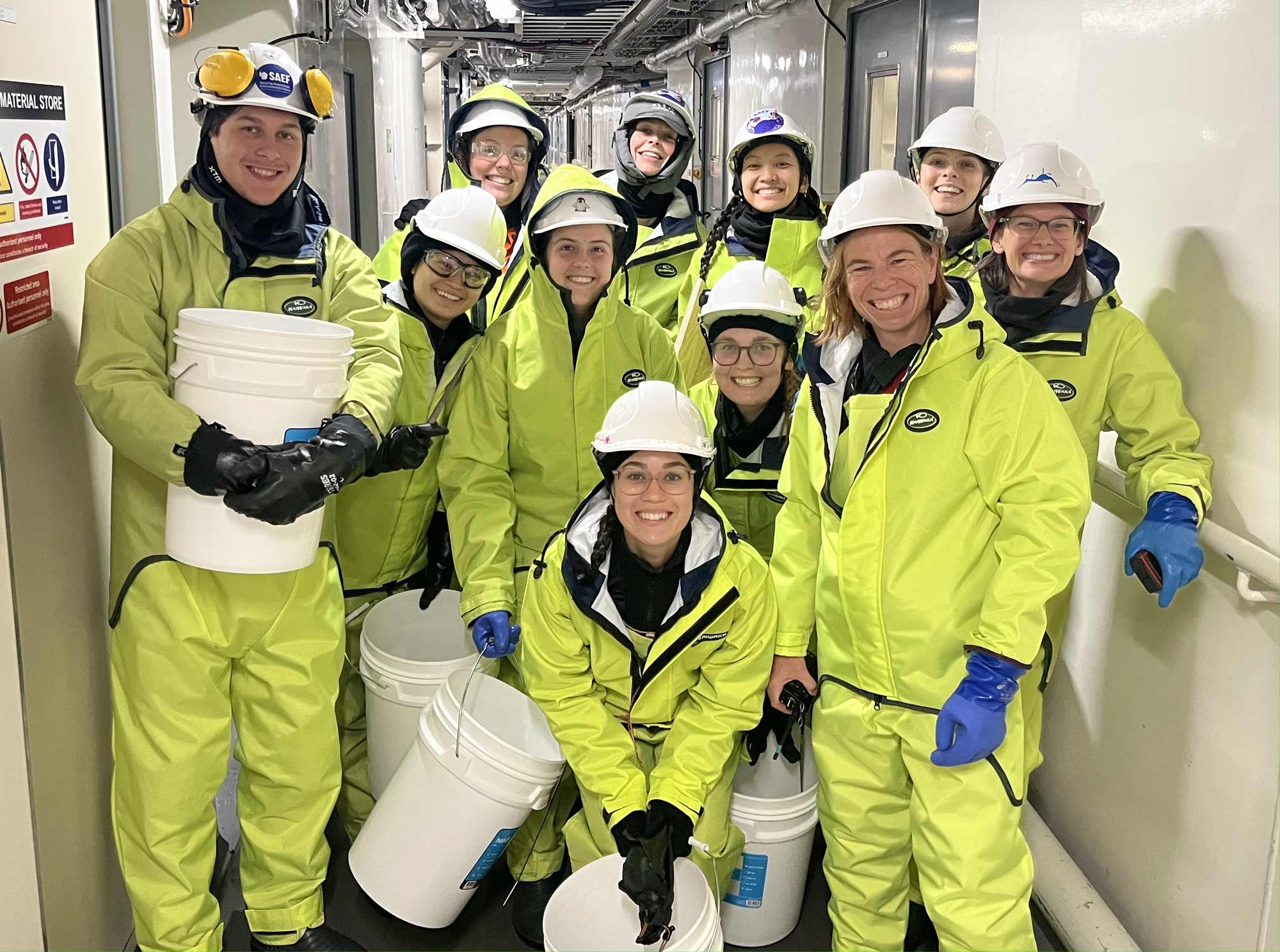 Eleven people in bright yellow survival suits, hard hats and surgical gloves stand in a corridor, many holding large white buckets