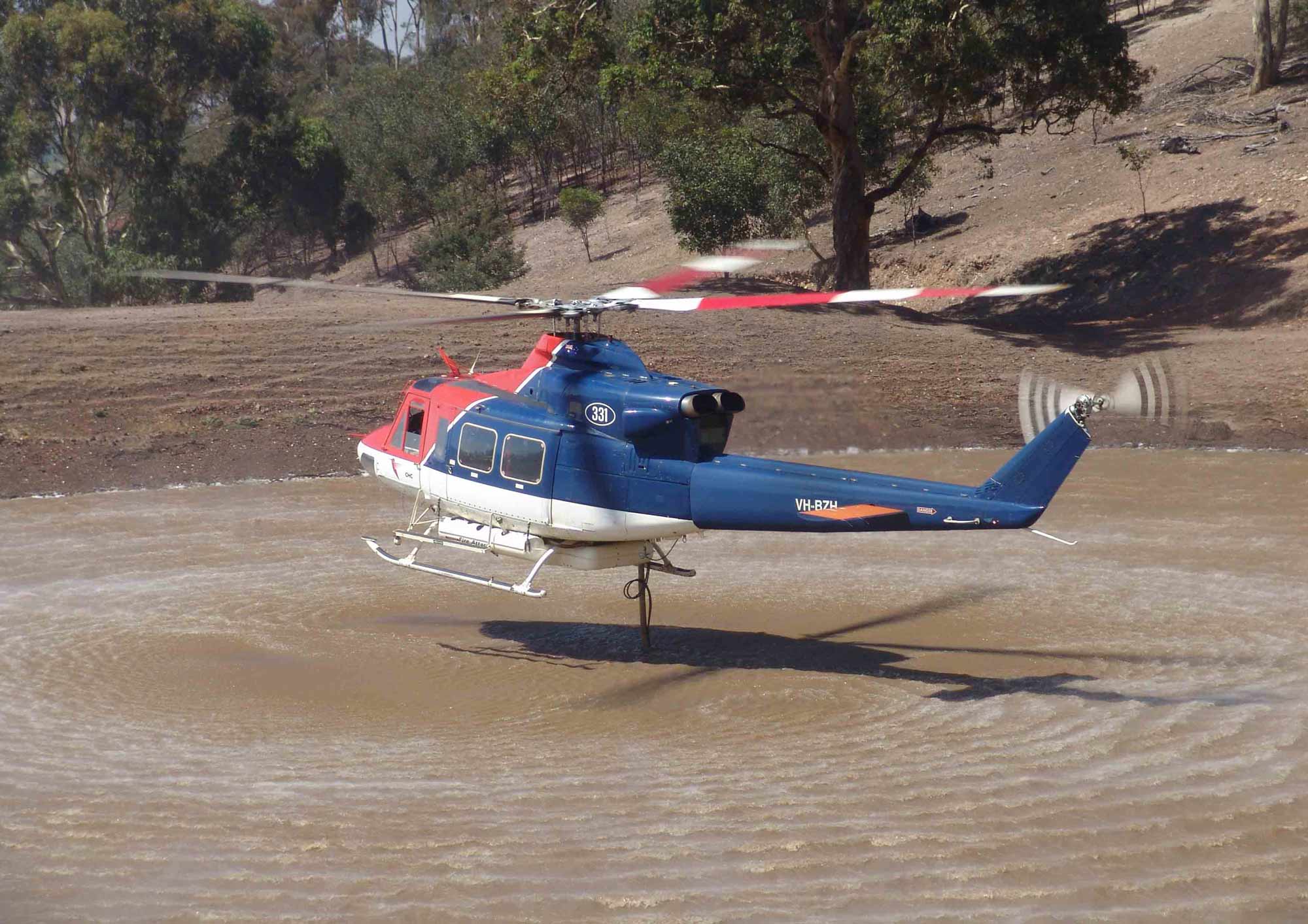Red and blue helicopter hovers over brown waterbody creating pressure ripples