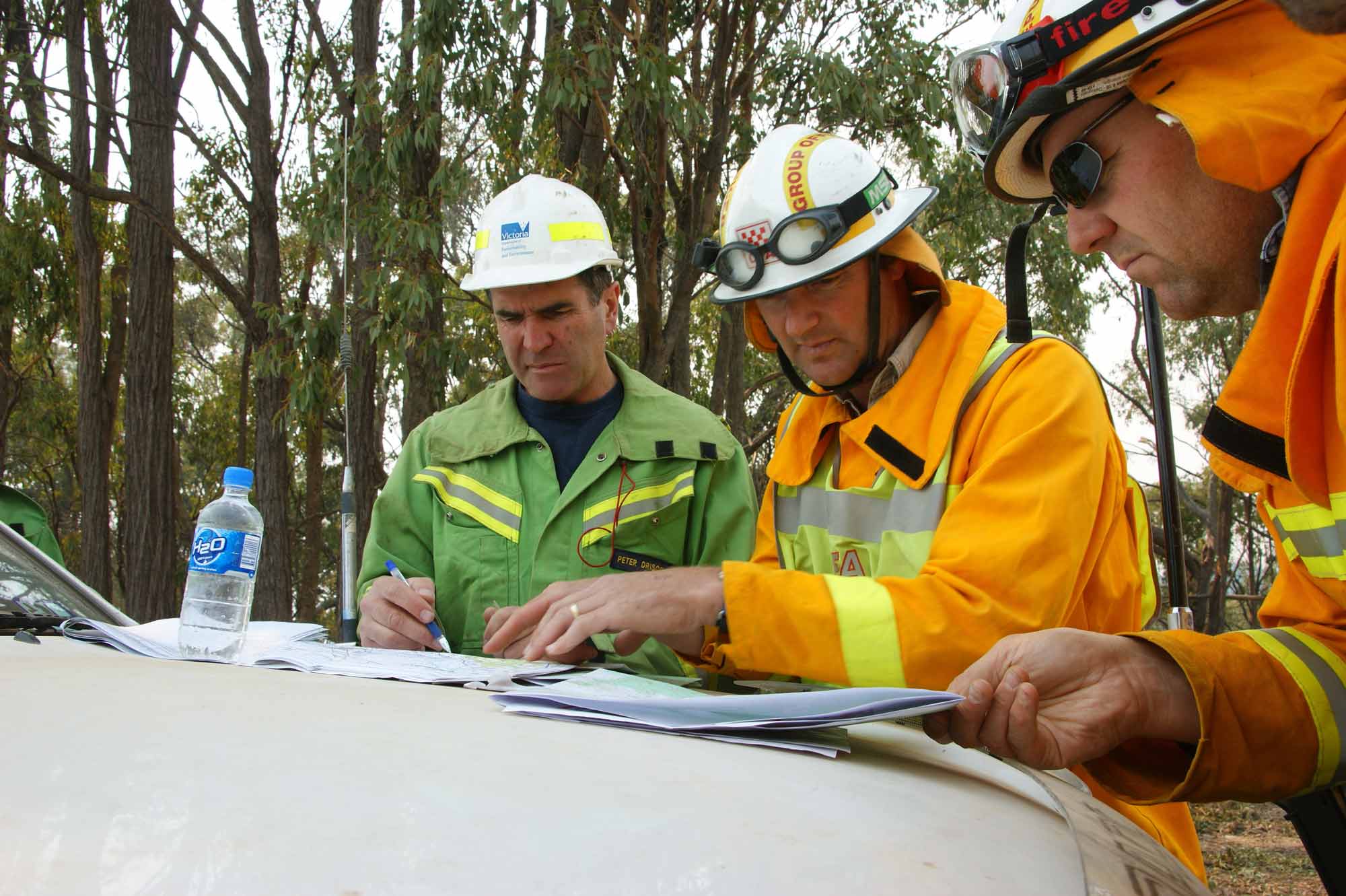 Three men in yellow and green emergency uniforms and hardhats collaborate on boot of a white vehicle